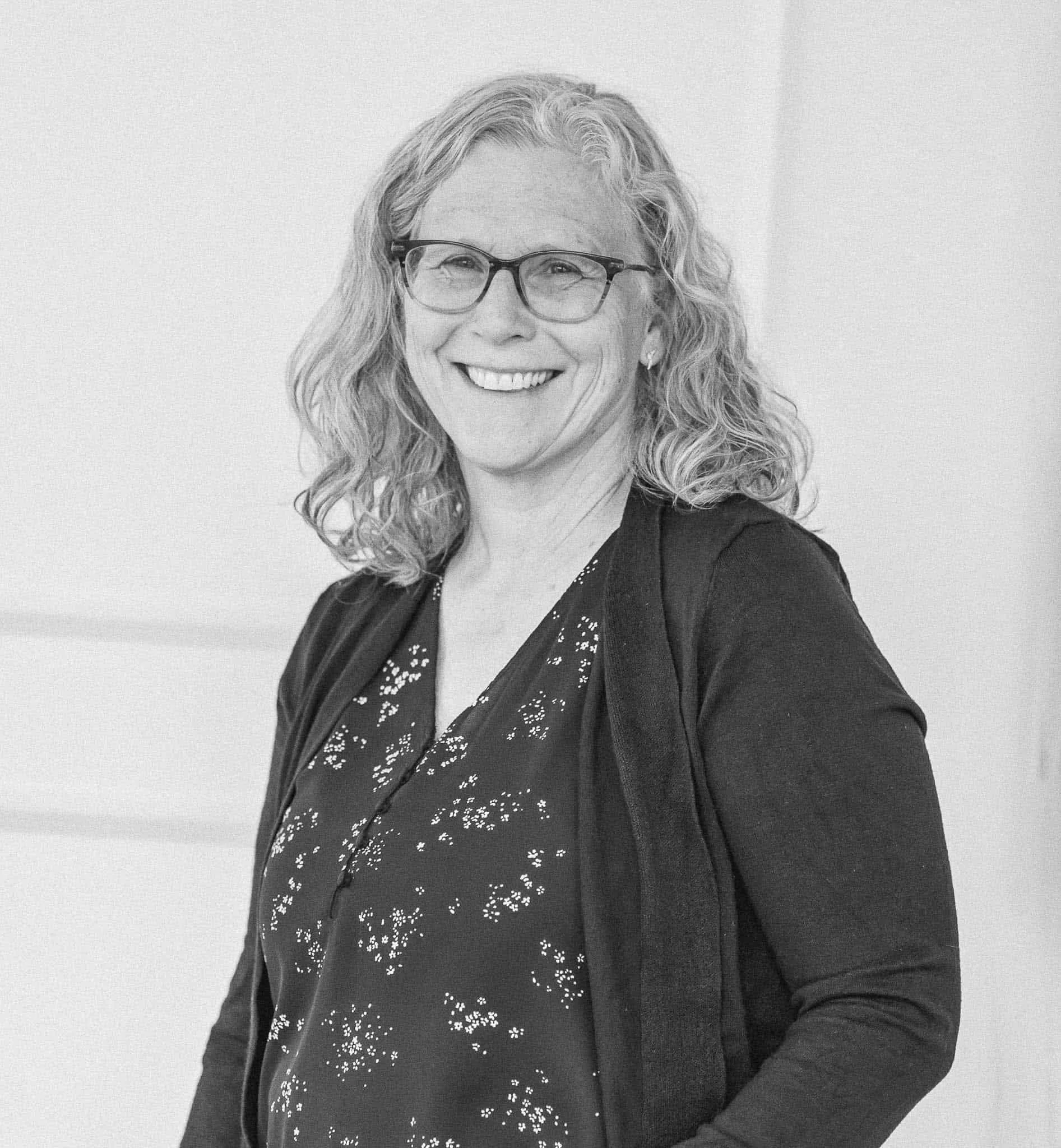 A smiling woman with curly hair, glasses, and earrings wearing a dark blazer and patterned blouse.