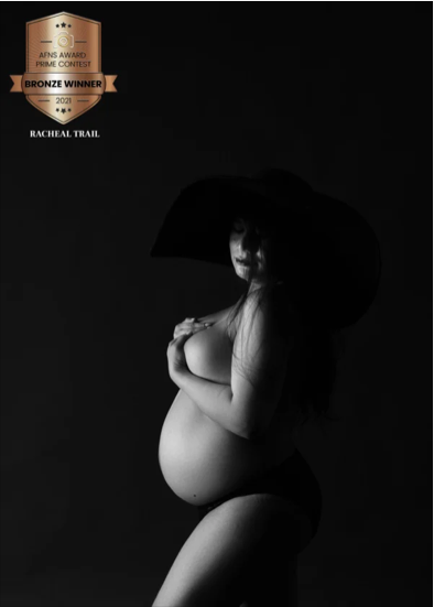 Black and white photograph of a pregnant woman wearing a large hat, with her hand resting on her belly, against a dark background. An award badge for AFNS Award Prime Contest 2021 Bronze Winner is in the top left corner.