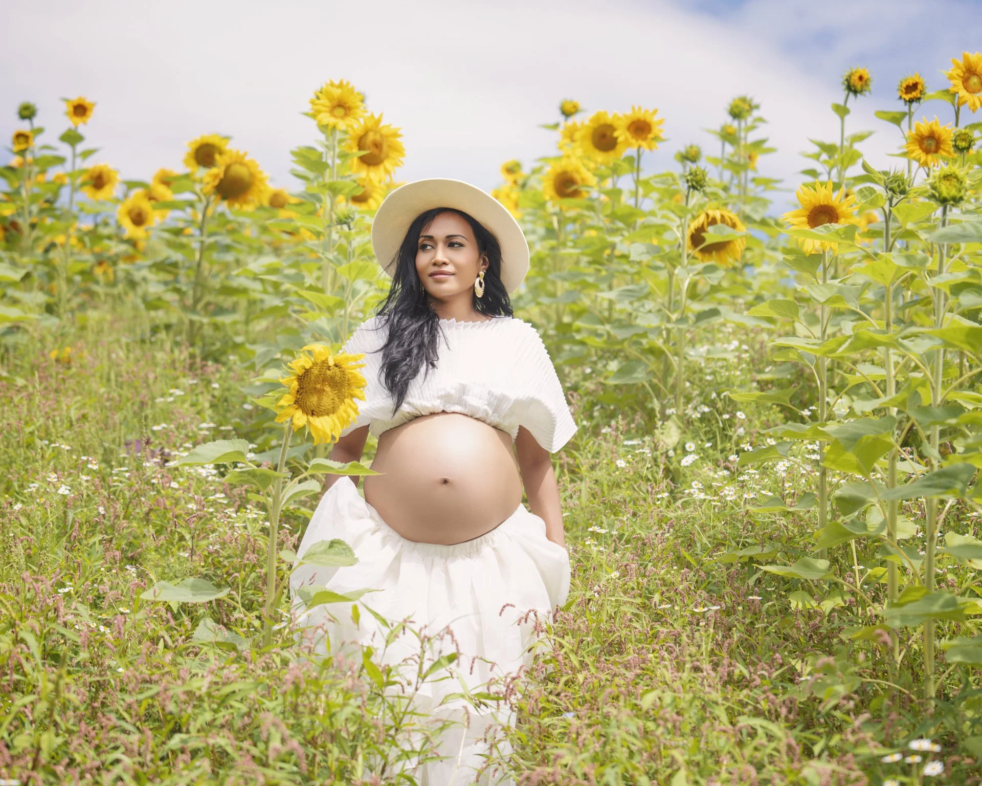 A pregnant woman with long dark hair, wearing a white hat, white top, and white skirt, stands in a sunflower field holding a sunflower, with a partly cloudy sky in the background.