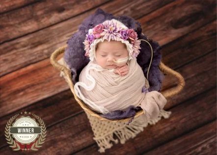 Young baby sleeping in a basket with a floral headband and wrapped in a blanket, on wooden floor.