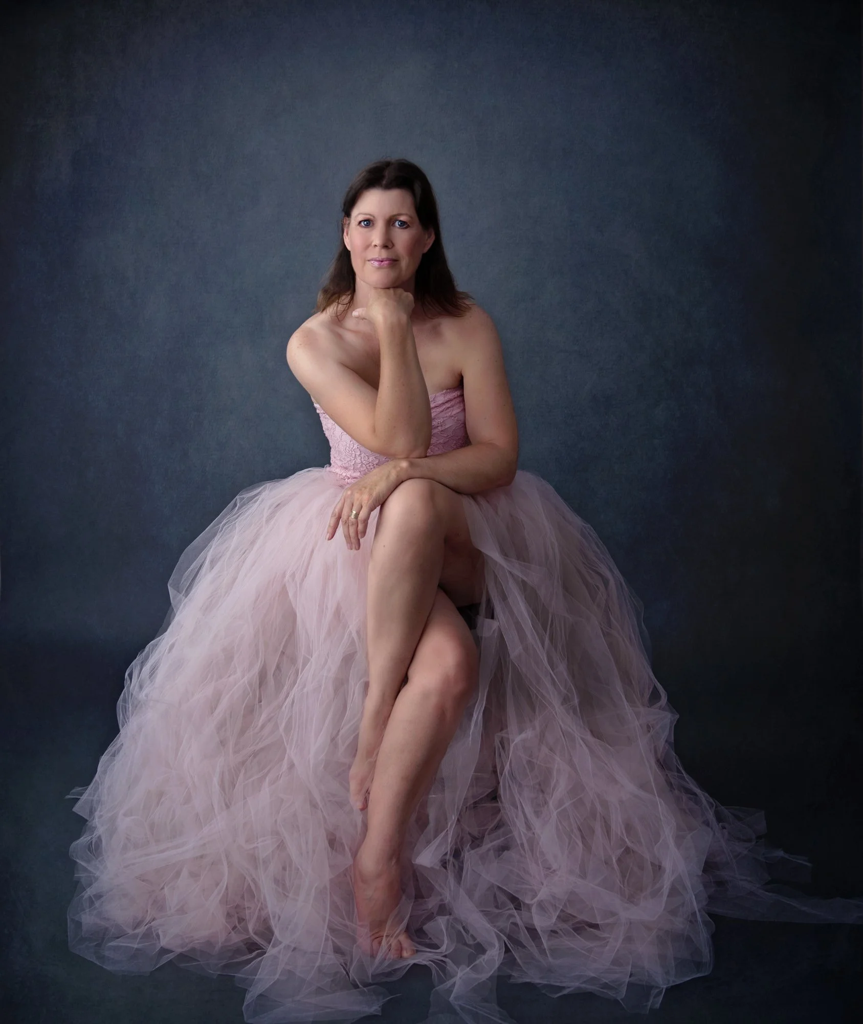 A woman in a pink strapless tulle gown sitting on a dark studio background, looking at the camera with her chin resting on her hand.
