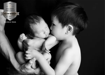 A young boy and a newborn baby sharing a tender moment, with the boy gently holding the baby and their faces close together, in black and white.