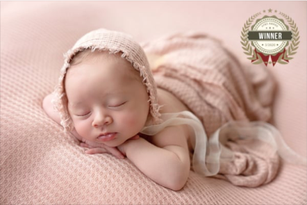 A sleeping baby lying on a pink textured blanket, wearing a knit hat and a light-colored blanket, with a medal badge in the top right corner that says "Winner".