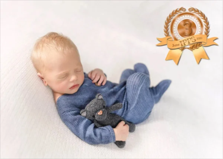 Young child sleeping on a white surface, holding a small black stuffed animal, with an award ribbon in the top right corner.