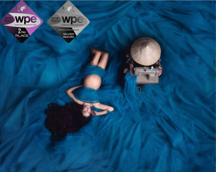 A woman in a blue dress lying on large blue fabric, surrounded by awards and a typewriter.