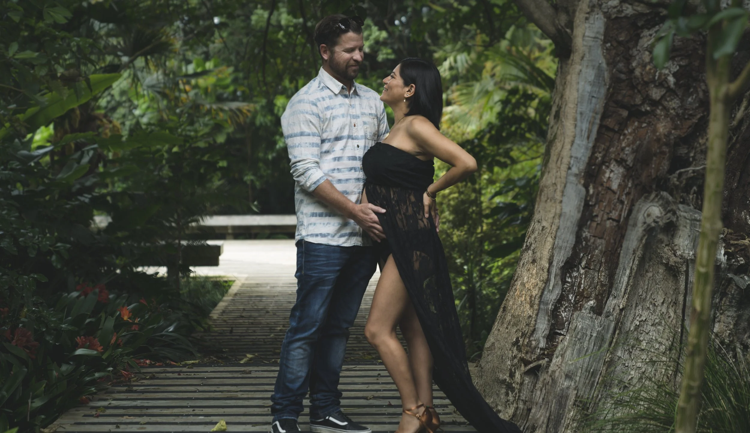 A man and woman standing on a wooden pathway in a lush green forest, gazing at each other, with the woman wearing a black dress and the man wearing a striped shirt and jeans.