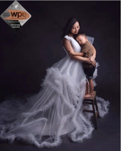 A woman wearing a long, flowing white dress holds a young child embracing her, standing on a small stool against a dark background. There is a WPE Awards logo in the top left corner.
