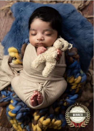 A sleeping baby wrapped in a beige blanket, holding a stuffed animal, lying on a colorful blanket with a blue pillow behind them.
