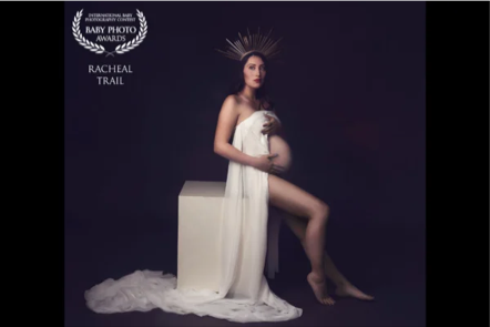 A woman dressed as a goddess, wearing a crown and a long flowing white gown, seated on a pedestal against a dark background.