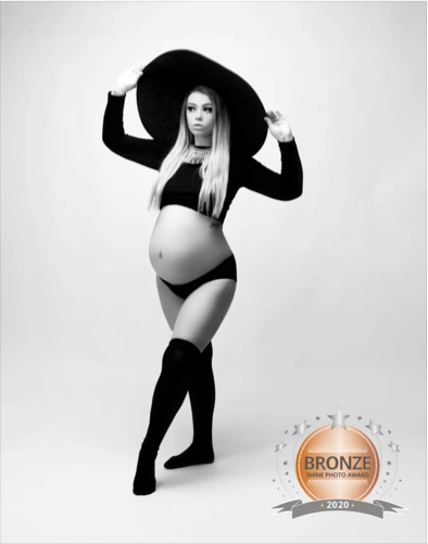 A woman with long blonde hair wearing a large sunhat, a black long-sleeve crop top, black underwear, and thigh-high stockings, posing against a plain background. She is holding the hat with both hands and has an award badge in the bottom right corner