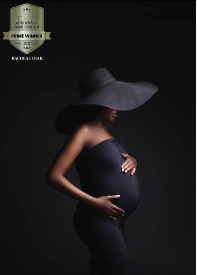 A pregnant woman wearing a large floppy hat and a strapless black dress, standing in profile against a dark background.