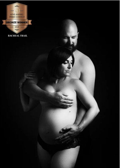 Black and white portrait of a pregnant woman with short dark hair and a bearded man. The man is holding the woman from behind, with one hand on her shoulder and the other on her stomach. The woman has her eyes closed and her hand resting on her belly