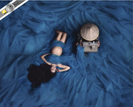 A woman with long dark hair laying on her back on a blue fabric surface, next to a small vintage typewriter and a round wooden tray.