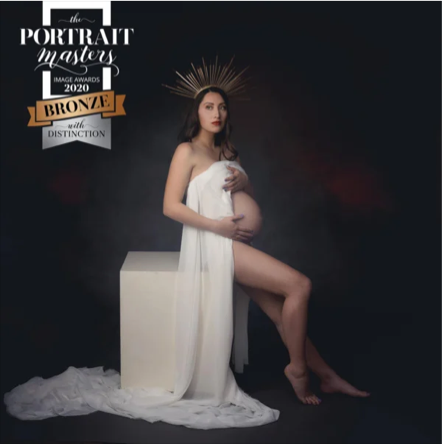 A pregnant woman sitting on a white block, wrapped in a white cloth, with a dark studio background and a burst of light behind her head, holding her belly. She is looking at the camera.