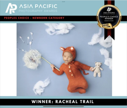 Cute baby dressed in a brown bear costume lying on a blue background, holding a dandelion puff, with scattered clouds and a teddy bear beside.