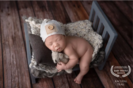 A sleeping baby lying on a small bed with pillows and blankets, wearing a knit cap and holding a plush toy, on a wooden floor.
