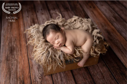A sleeping baby lying in a wooden box with a fluffy blanket, on a wooden floor.