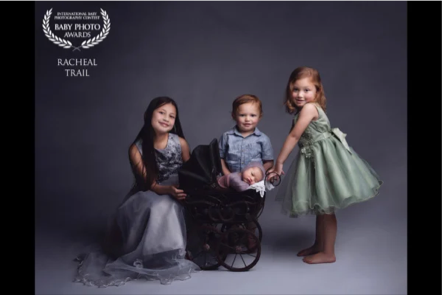 Three young children in formal attire posing for a portrait with a stroller against a dark background. The girl on the left wears a gray dress, the boy in the middle sits in the stroller, and the girl on the right wears a green dress.