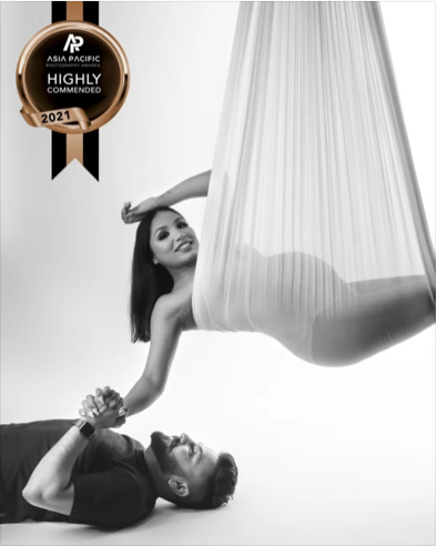 A woman is hanging in an aerial silk, smiling. A man lies on his back on the floor, holding her hand. The image is in black and white with a badge indicating a 2021 Asia Pacific Highly Commended award.