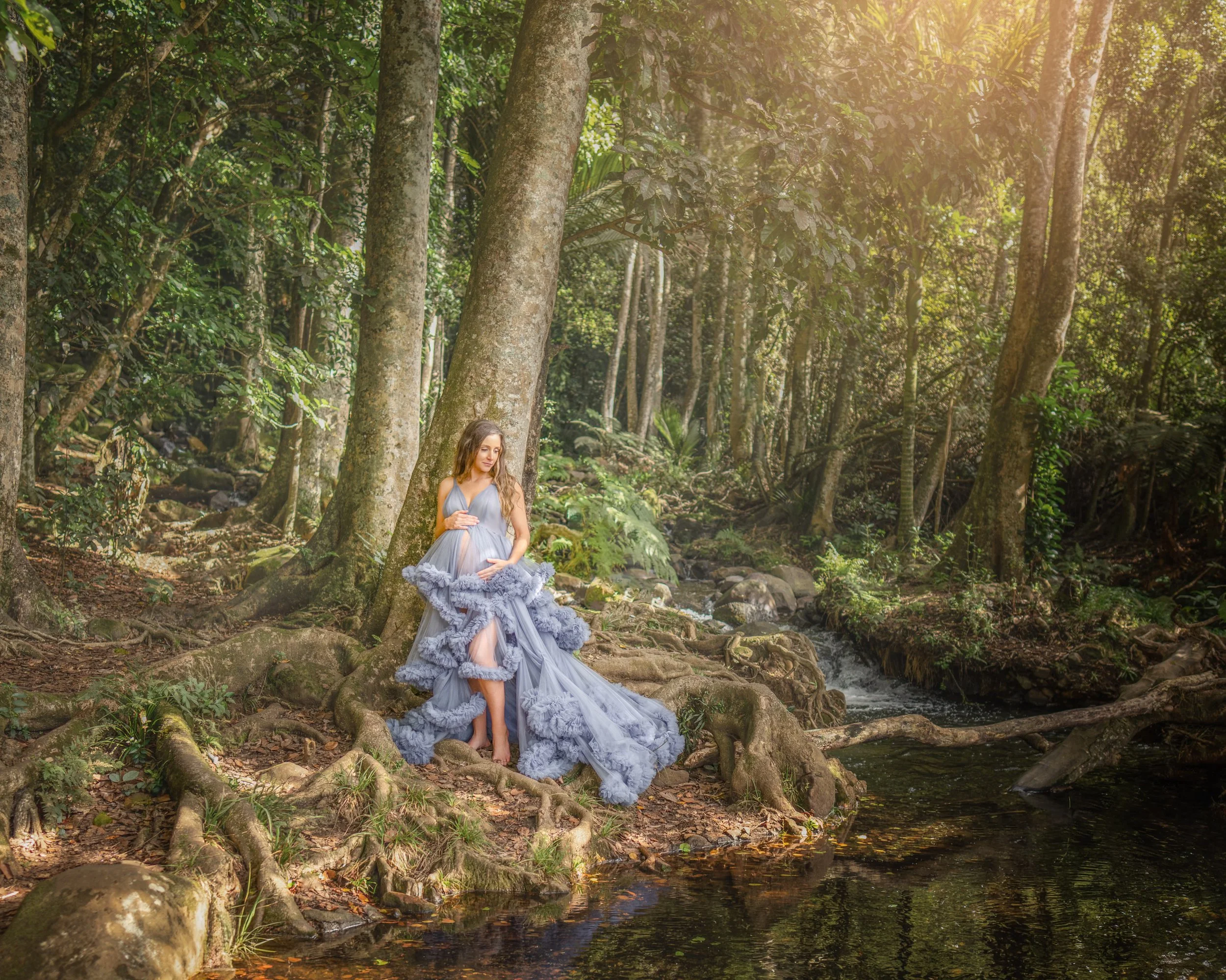 Pregnant woman in a flowing blue gown beside a forest stream during an outdoor Auckland maternity photography session.