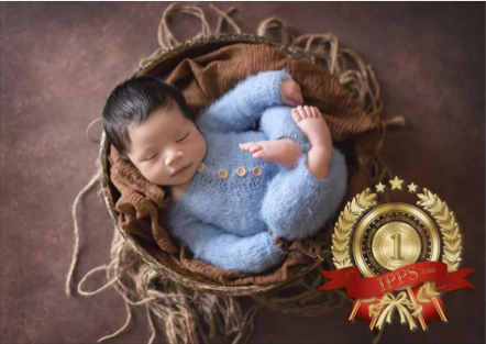 A newborn baby sleeping in a wicker basket, wrapped in a light blue fuzzy blanket, with a medal and ribbon indicating first place.