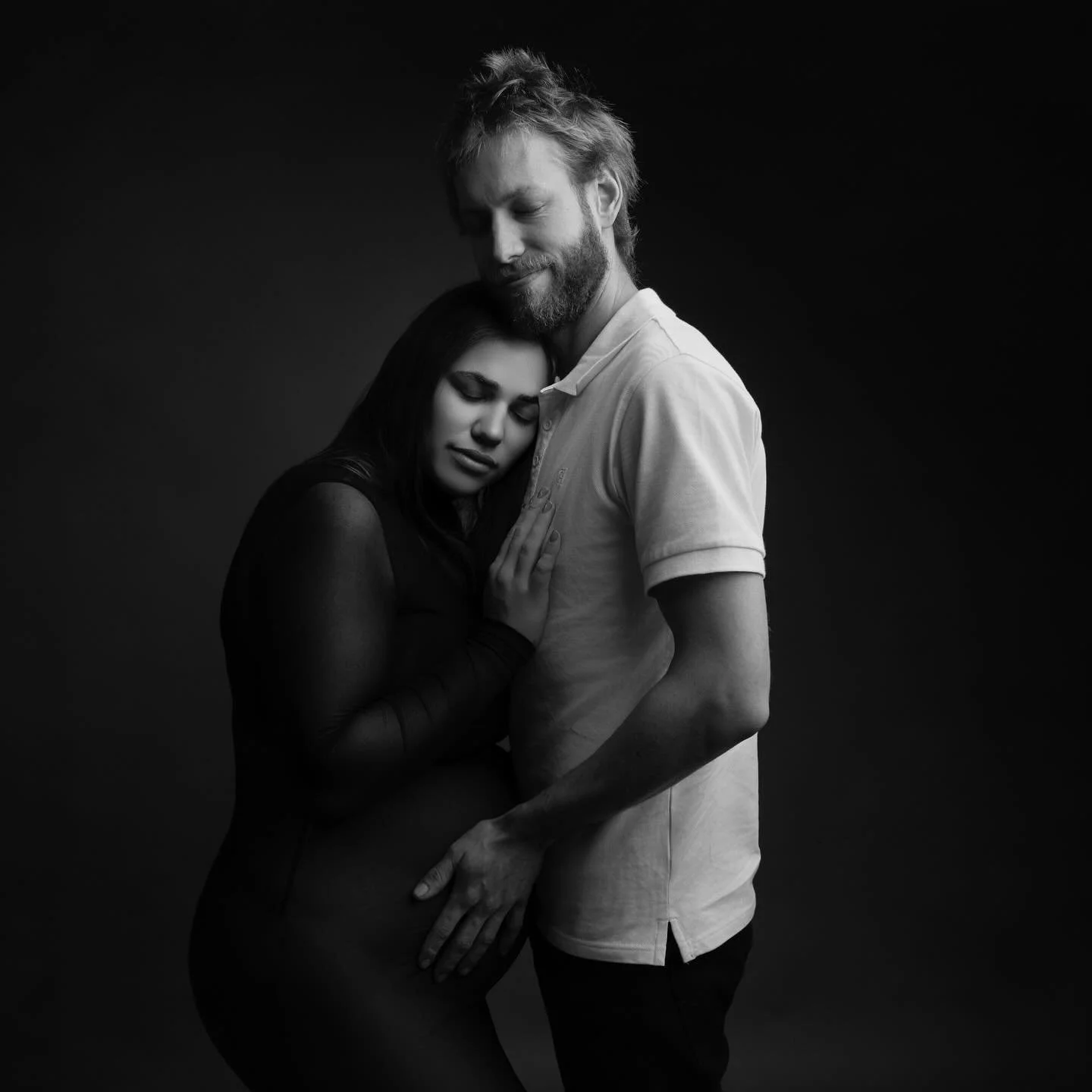 A black-and-white photo of a man and woman embracing; the woman rests her head on the man's chest with eyes closed, and the man gently holds her with a serene expression.