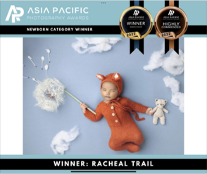 A baby dressed in a brown knit outfit with bear ears, lying on a blue background with cloud cutouts and holding a fluffy white umbrella shaped like a cloud, alongside a small white teddy bear. The image is part of the Asia Pacific Photography Awards,
