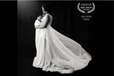 Black and white photo of a pregnant woman wearing a flowing gown, holding her belly, with a backdrop and text indicating a baby photo award.