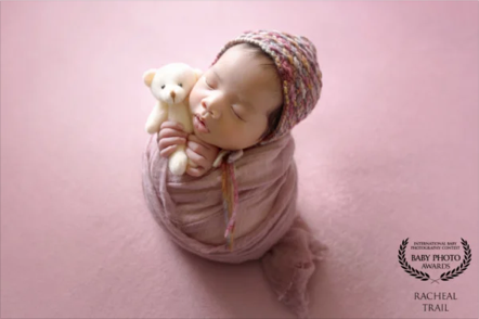 Baby wrapped in pink blanket, with head resting on a teddy bear, against a pink background.