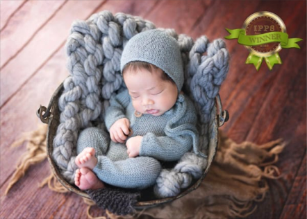 A sleeping baby in a knitted gray hat and sweater, nestled in a cozy, chunky gray blanket inside a round basket on a wooden floor, with a gold and green award badge in the top right corner.