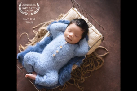 Newborn baby sleeping on a small bed with a blue fuzzy onesie, in a cozy setup with a brown background.