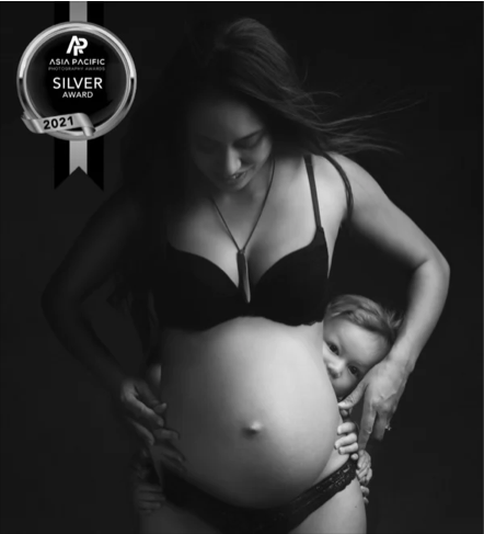 Black and white photo of a pregnant woman with a young child hugging her belly, smiling, with an Asia Pacific Silver Award badge from 2021.