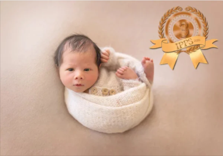 Baby lying in a white knitted blanket on a beige surface, looking at the camera, with a gold badge featuring a '7' and ribbon in the upper right corner.