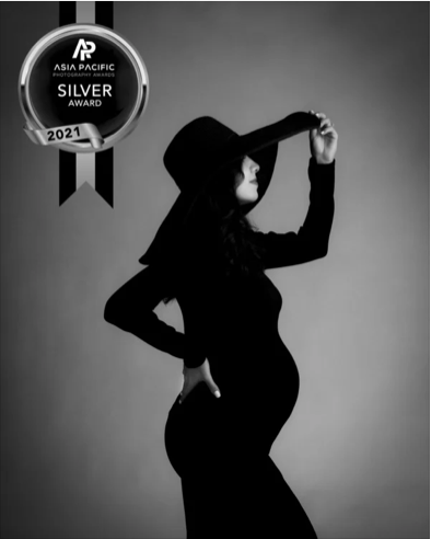 Black and white side profile of a pregnant woman wearing a large hat and a long dress, holding her hat with one hand and resting her other hand on her hip, with an Asia Pacific Photography Award Silver Award badge from 2021 in the top left corner.
