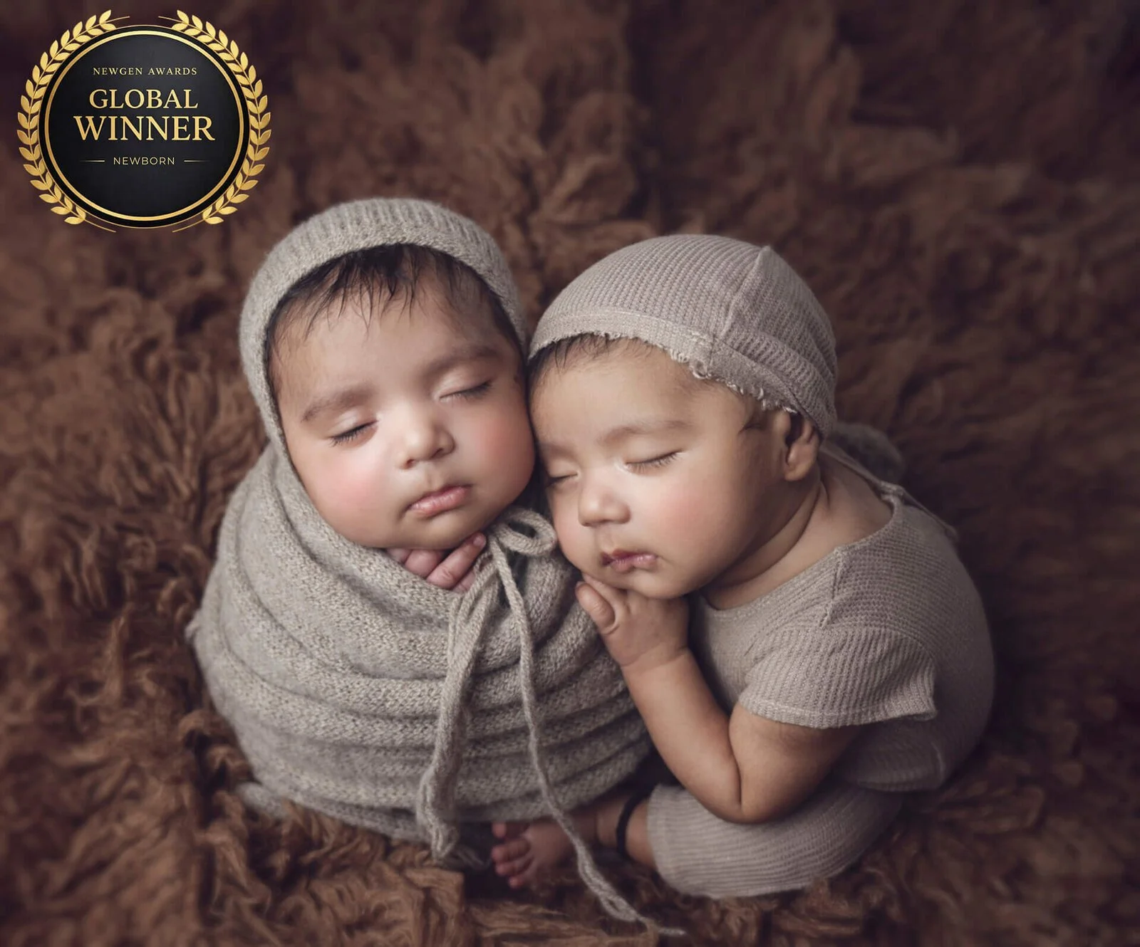 Two sleeping babies, wrapped in beige cloth and wearing beige hats, lying on a brown furry blanket, with the baby on the right holding the other baby's hand. A black and gold badge in the top left corner reads 'Newgen Awards Global Winner Newborn.' 