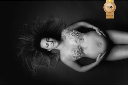 A woman with long dark hair lying on her back on a dark surface, wearing a revealing outfit and displaying tattoos on her chest.