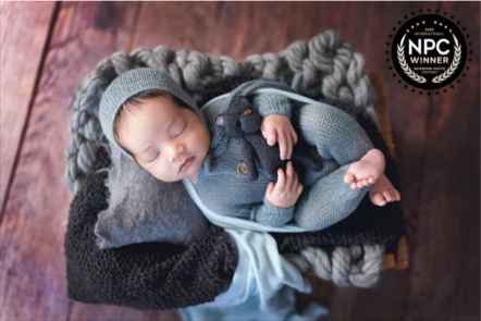 A sleeping baby in a gray beanie, wrapped in a blue blanket, resting on a gray pillow, surrounded by knitted blankets on a wooden surface, with an NPC winner badge in the top right corner.