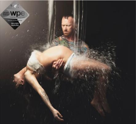 A man with wet hair holding a person upside down under a waterfall, with water splashing around them.