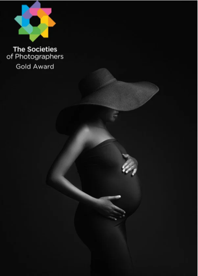 Black and white photo of a pregnant woman wearing a large sun hat, holding her belly and resting her other hand on her side. There is a logo in the top left corner that says 'The Societies of Photographers' with a gold award label.