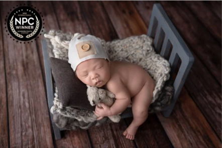A sleeping baby with a white hat featuring a bear face, lying on a small bed with a gray headboard and soft blanket, on a wooden floor.