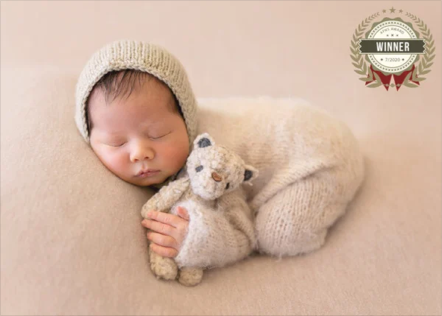 A sleeping newborn baby wearing a beige knit hat, cuddling a small teddy bear while lying on a beige blanket.