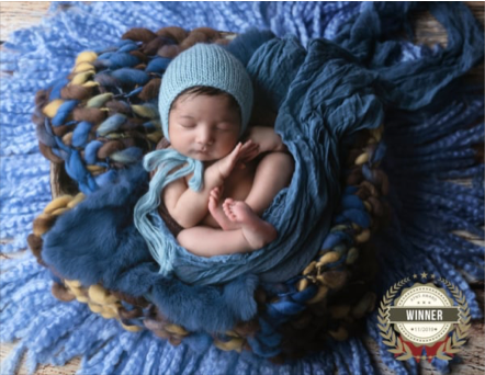 A sleeping newborn baby wrapped in a blue blanket and wearing a blue knit hat, lying in a dark brown woven basket on a textured blue blanket.