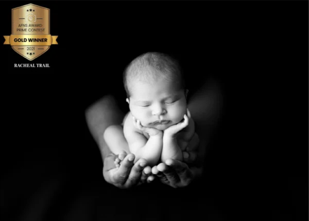 Black and white photo of a sleeping baby cradled in adult hands with an award badge in the top left corner