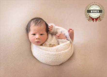 Baby lying in a white, knitted basket with a beige background and a winner badge in the top right corner.