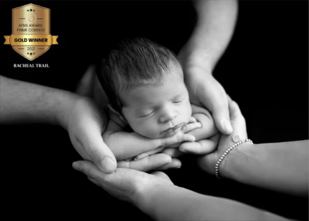 Black and white photo of a sleeping newborn being gently held by multiple hands, with a badge indicating a Gold Winner award for Rachel Trail.
