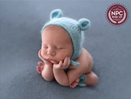 Baby wearing a blue knit hat with bear ears, resting chin on hands, on a soft gray surface.