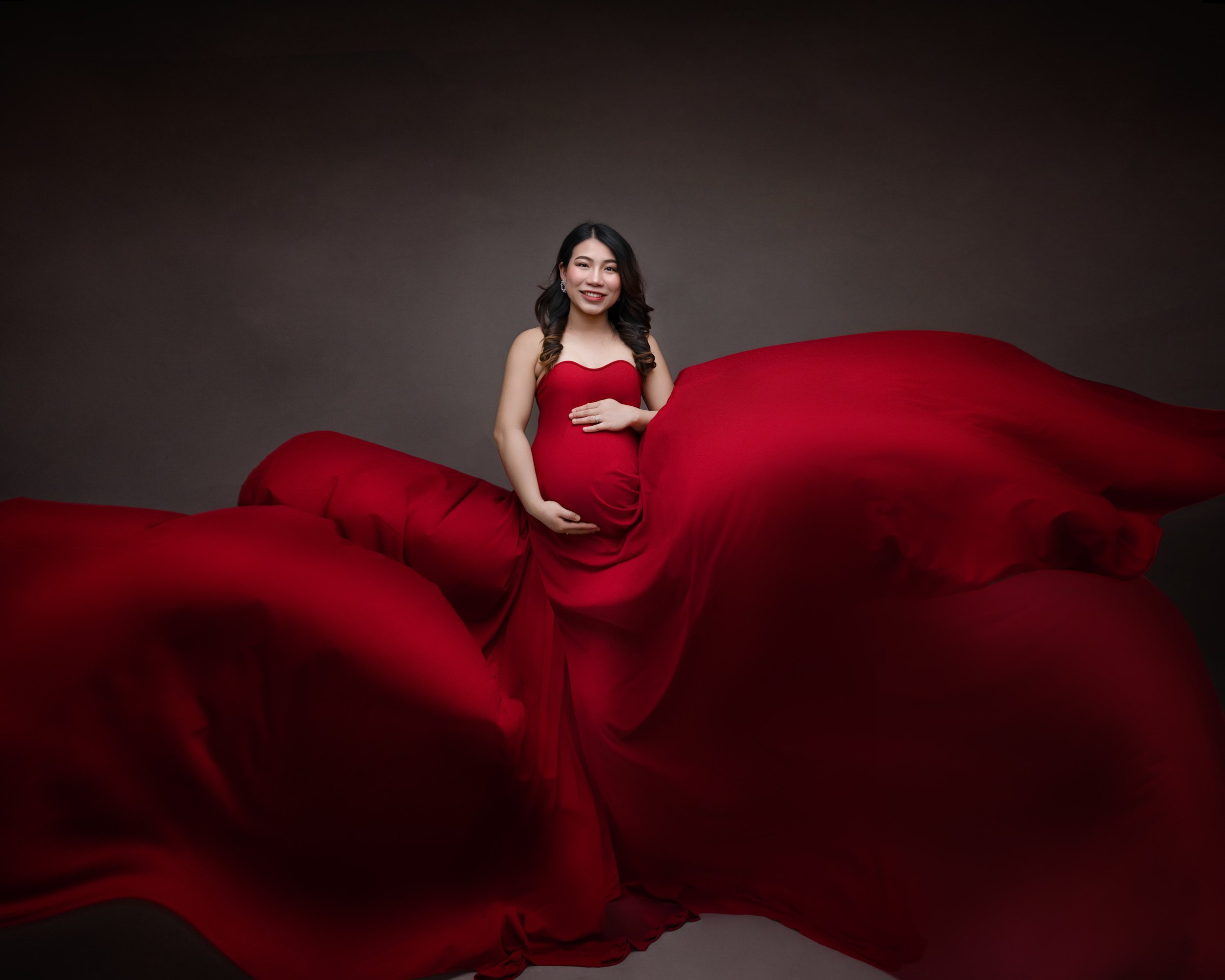 Auckland maternity photography portrait of an expectant mum in a flowing red gown with dramatic fabric movement in a studio setting.