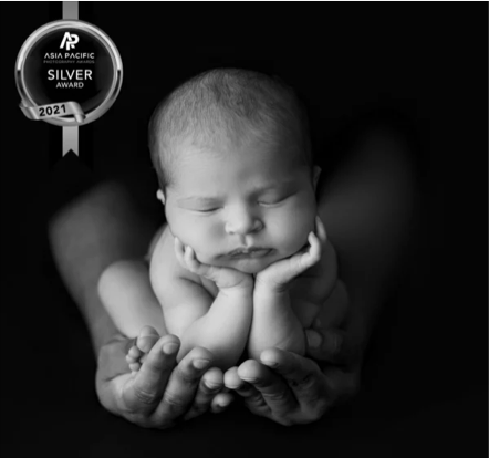 Black and white photo of a sleeping newborn baby with chin resting on hands, cradled in adult hands, with an Asia Pacific Silver Award badge in the top left corner.