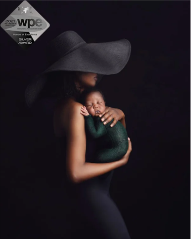 A woman wearing a large black wide-brimmed hat holding a sleeping baby wrapped in a green blanket against a dark background.
