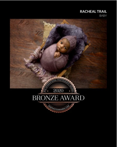 Photograph of a baby doll wrapped in a purple blanket, lying in a wooden cradle on a dark wooden floor, with a golden pillow behind. Text overlay indicates Racheal Trail Baby, 2020 Bronze Award from the International Photography Awards.
